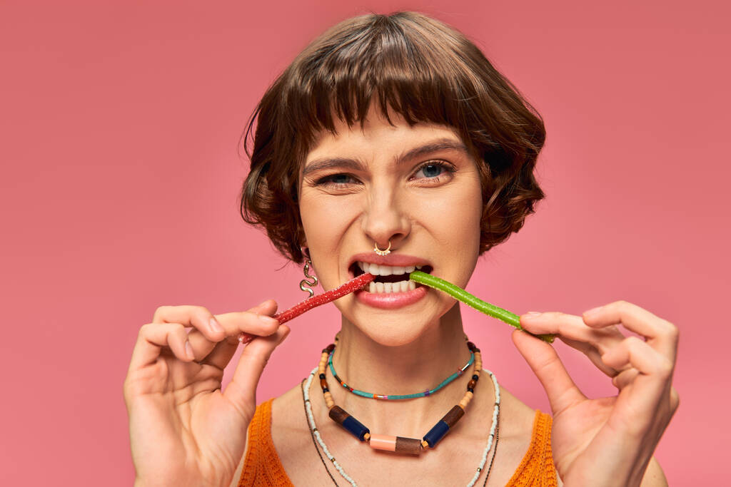 funny pierced girl in 20s biting two different flavors of sweet and sour candies on pink backdrop - Photo, Image