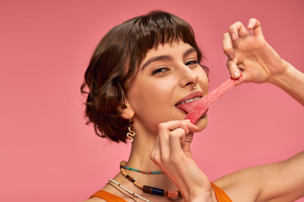 весела і молода жінка з пронизливим носом лиже солодку і кислу смужку цукерок на рожевому фоні - Фото, зображення