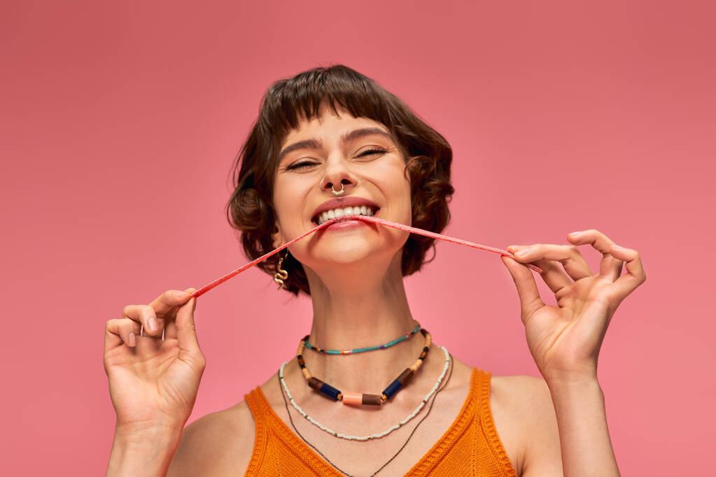 Восхитительная и молодая женщина с пирсингом носа кусает сладкие и кислые конфеты полоску на розовом фоне - Фото, изображение