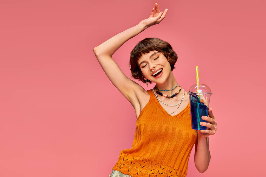 ピンクの頭の上に爽やかな夏の飲み物を保持する短いブルネット髪の興奮した若い女性 - 写真・画像