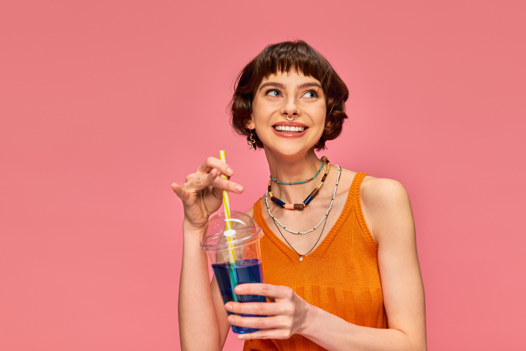 pleased young woman with short brunette hair holding refreshing summer drink on pink backdrop - Photo, Image