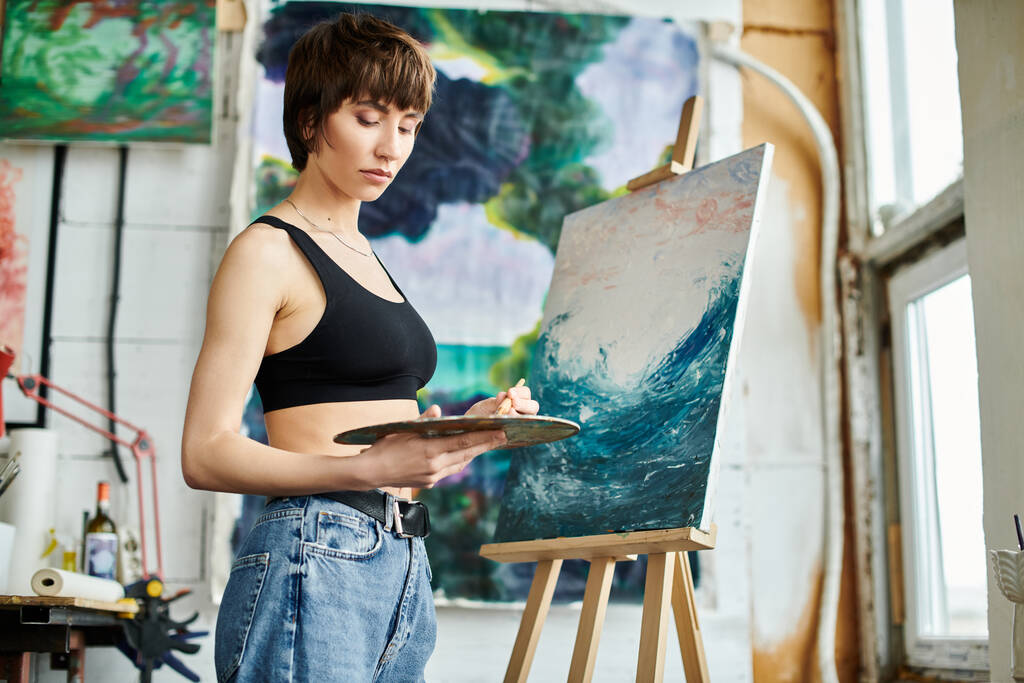 Una mujer en una camiseta negra se centra en la pintura en un caballete. - Foto, Imagen