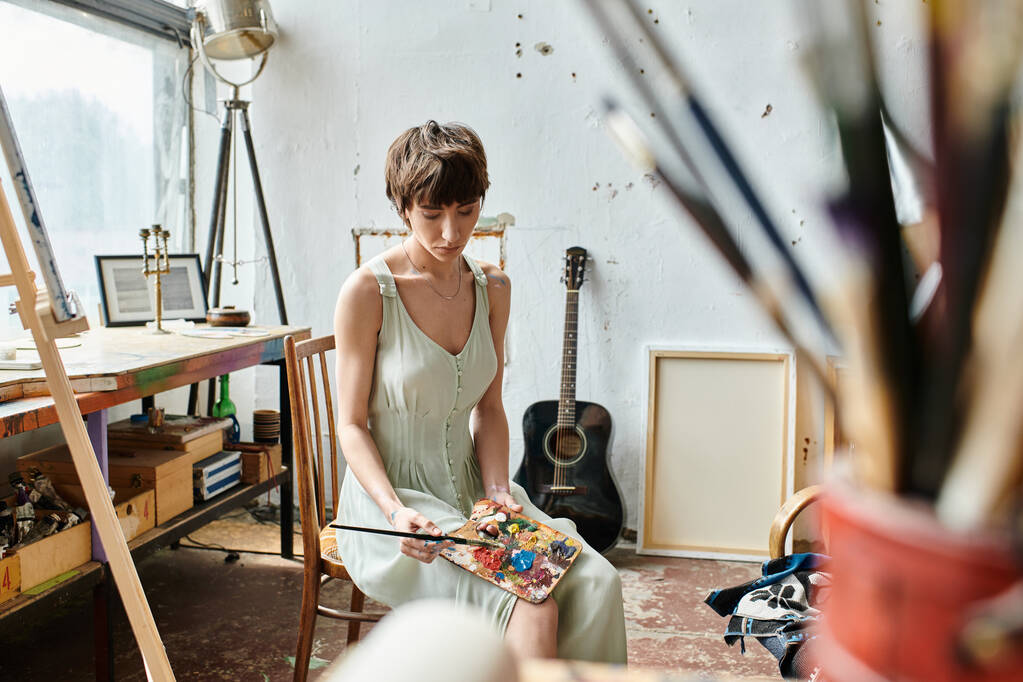 Kobieta siedzi w pokoju z gitarą, trzymając pędzel i paletę. - Zdjęcie, obraz