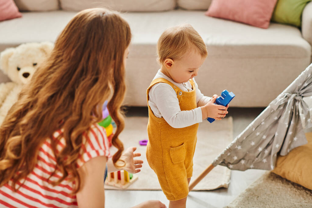Un niño pequeño, acompañado por su madre, explora alegremente la sala de estar con un juguete colorido. - Foto, Imagen