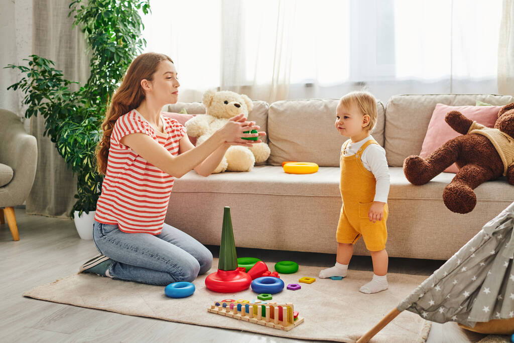 Μια νεαρή μητέρα ασχολείται παιχνιδιάρικα με την κόρη της εν μέσω παιχνιδιών σε ένα άνετο σαλόνι. - Φωτογραφία, εικόνα