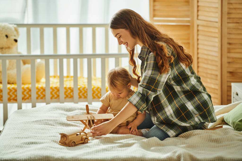 Una giovane madre gioca con gioia con sua figlia su un letto accogliente, condividendo sorrisi e risate in una scena commovente.. - Foto, immagini