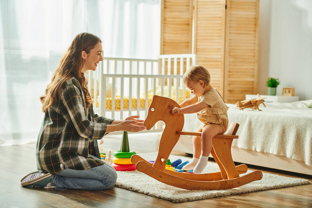Uma jovem mãe brinca alegremente com sua filha criança em um cavalo de balanço de madeira, criando doces lembranças em casa.. - Foto, Imagem