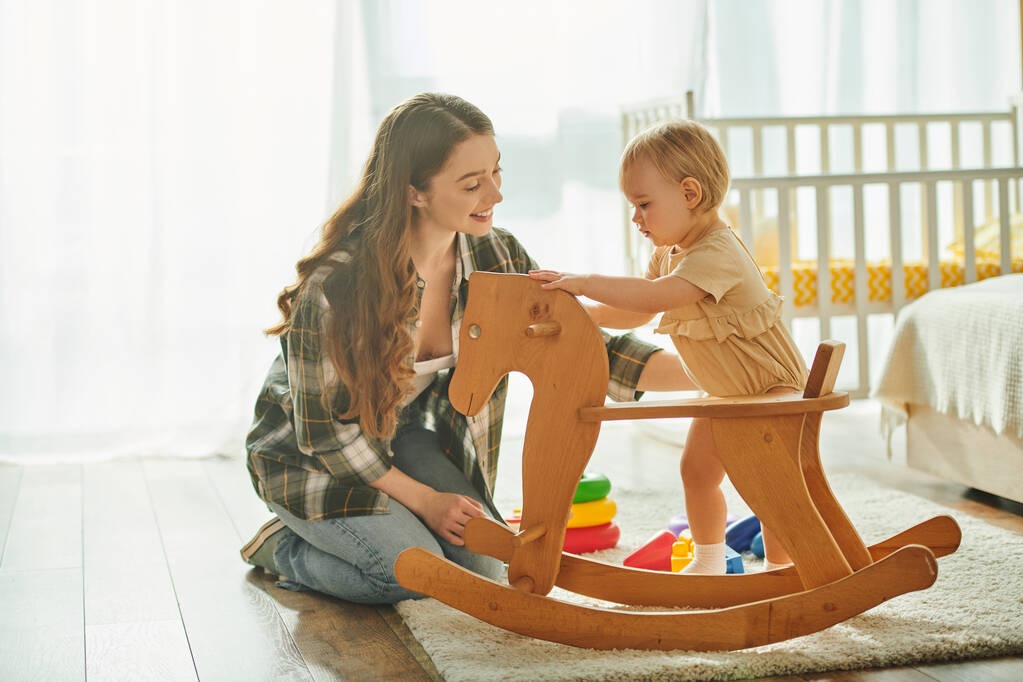 Młoda matka bawi się z córką na drewnianym koniu w przytulnym domu.. - Zdjęcie, obraz
