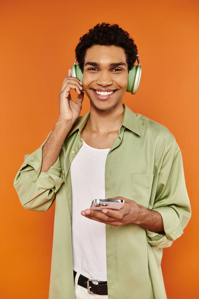 vreugdevolle stijlvolle Afrikaans-Amerikaanse man met een koptelefoon die zijn smartphone vasthoudt en lacht naar de camera - Foto, afbeelding