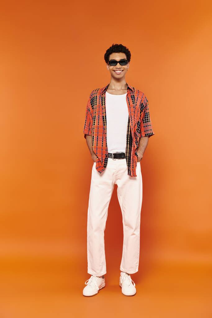 vreugdevolle jonge Afrikaanse Amerikaanse man in levendige kledij met een zonnebril poseren op oranje achtergrond - Foto, afbeelding