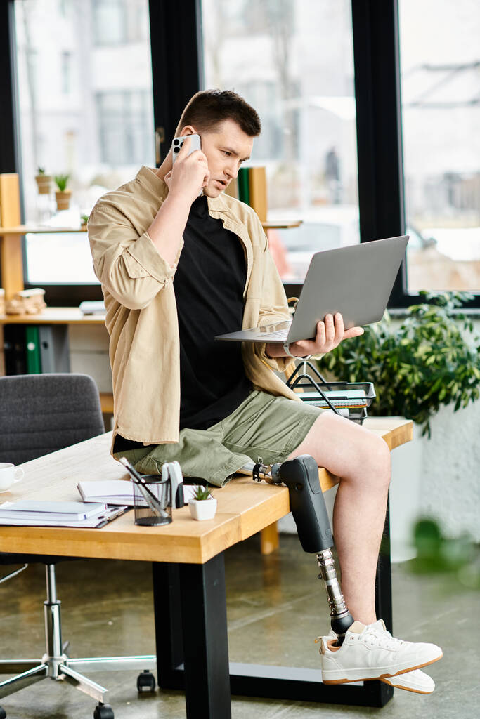 Un homme d'affaires avec une prothèse de jambe s'assoit à une table, utilisant un ordinateur portable et parlant sur un téléphone portable. - Photo, image