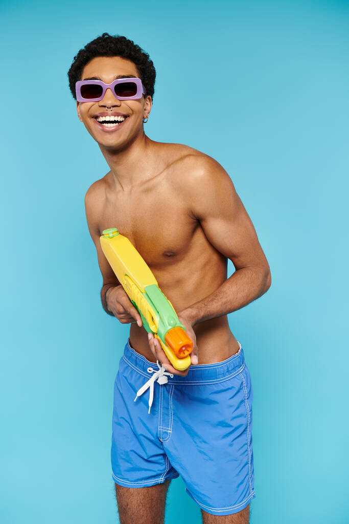 optimistischer junger afrikanisch-amerikanischer Mann in Badehose mit stylischer Sonnenbrille posiert mit Wasserpistole - Foto, Bild