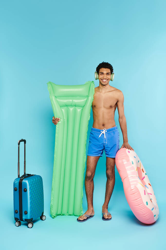 hombre americano africano alegre con los auriculares con el colchón de aire y la dona inflable al lado de la maleta - Foto, imagen