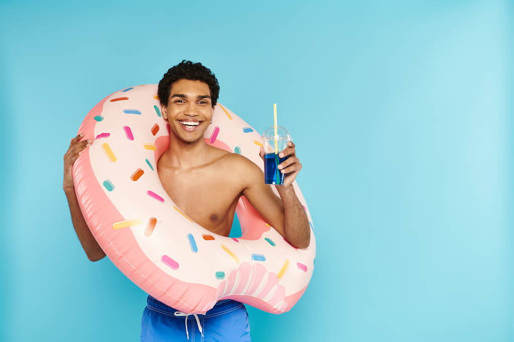 alegre afroamericano hombre con donut inflable disfrutando refrescante cóctel sobre fondo azul - Foto, Imagen
