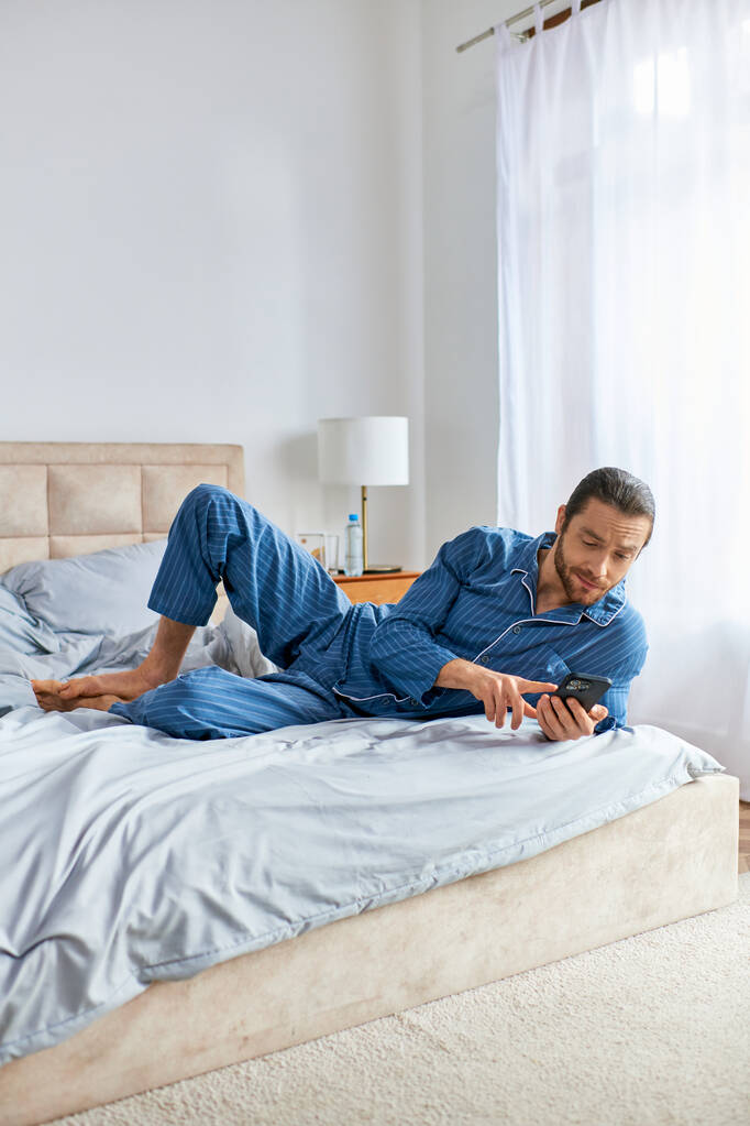 Un uomo pratica pacificamente yoga su un letto mentre è assorto nel suo cellulare. - Foto, immagini