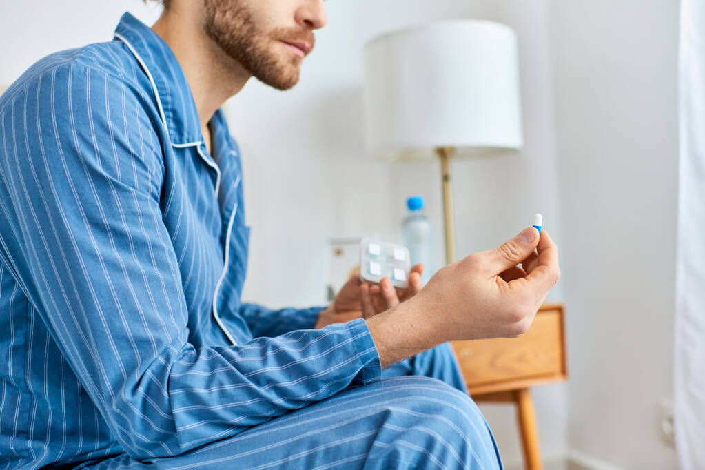 Un hombre sentado en una cama, tomando pastillas en un ambiente acogedor por la mañana. - Foto, imagen