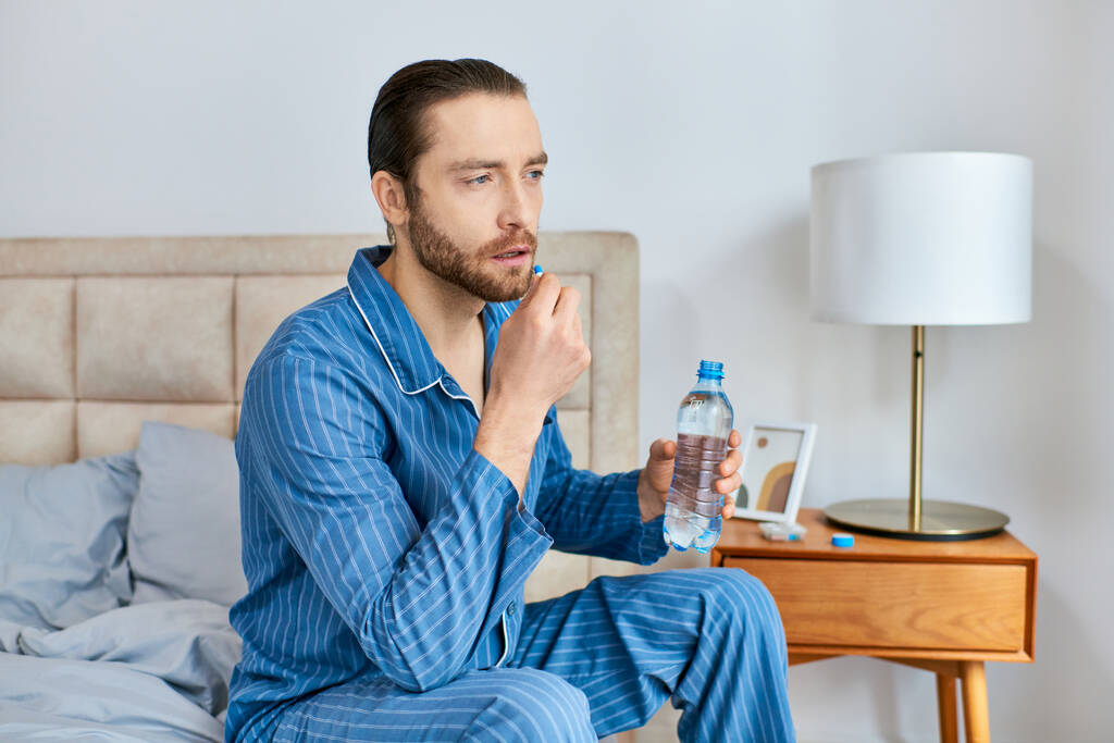 Człowiek siedzący na łóżku, trzymający butelkę wody i pigułkę, głęboko w myślach. - Zdjęcie, obraz