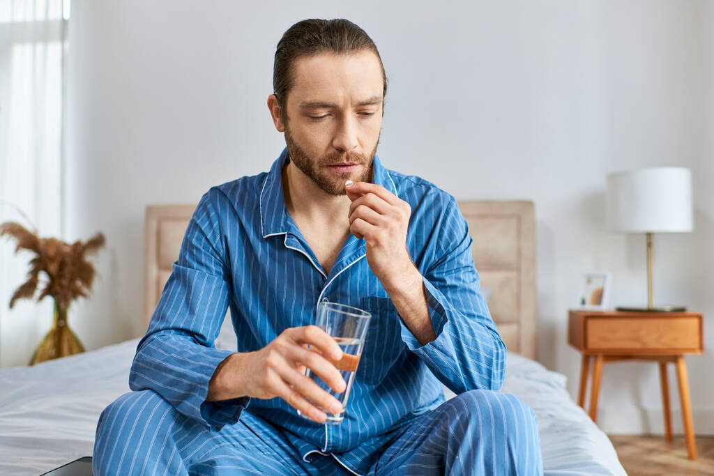 Um homem sentado em uma cama, bebendo pacificamente um copo de água com pílula. - Foto, Imagem