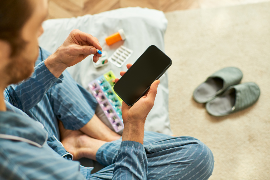 Een man die op de vloer zit houdt zich bezig met zijn smartphone en pillen. - Foto, afbeelding