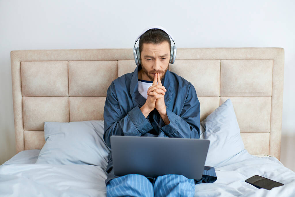 Un uomo seduto su un letto, con le cuffie, che usa un portatile. - Foto, immagini