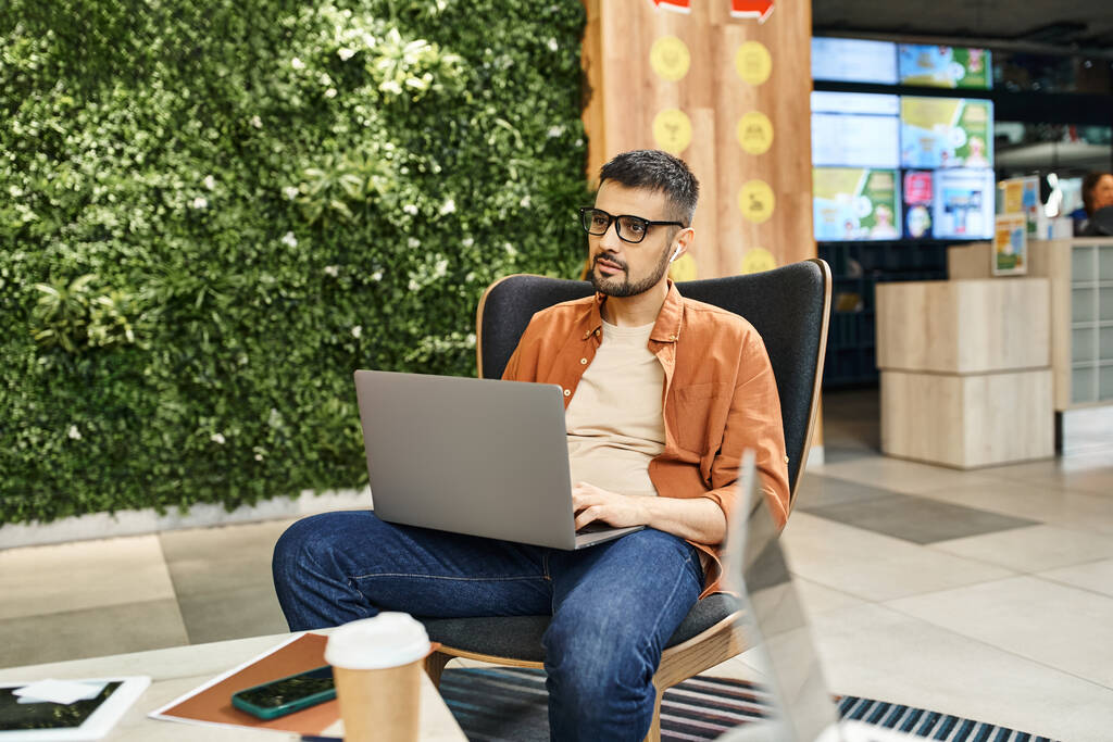 Een man in een stoel, ondergedompeld in zijn laptop, omgeven door de drukte van een co-working ruimte waar startup teamleden samenwerken. - Foto, afbeelding