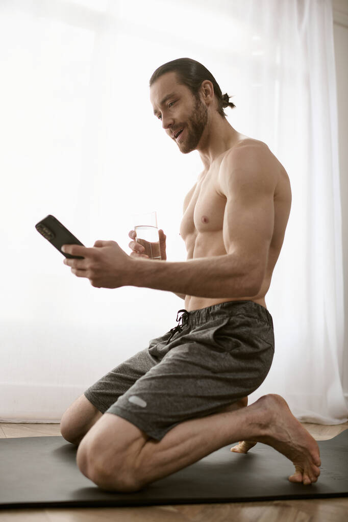 Ένας ημίγυμνος άντρας κάθεται σε ένα στρώμα γιόγκα, κρατώντας ένα κινητό τηλέφωνο, που ασχολούνται με το πρωινό διαλογισμό. - Φωτογραφία, εικόνα