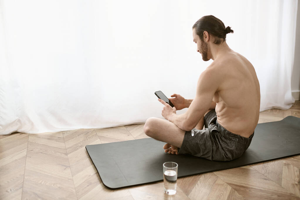 Um homem sentado em um tapete de ioga, usando um telefone celular. - Foto, Imagem