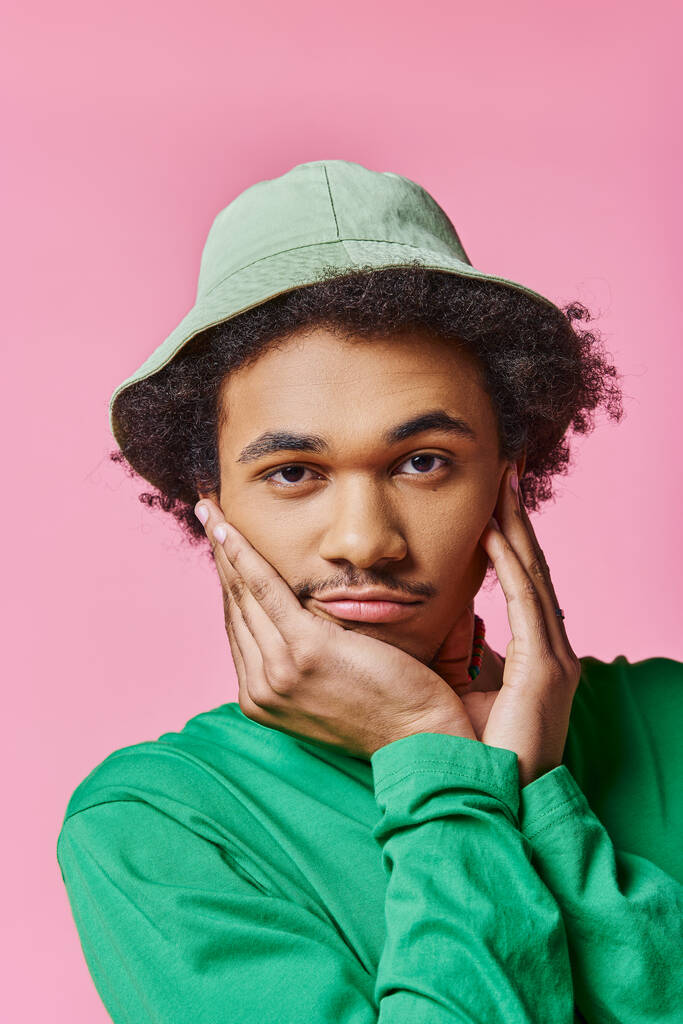 Скучающий афроамериканец в зеленой рубашке и шляпе на розовом фоне источает очарование и эмоции. - Фото, изображение