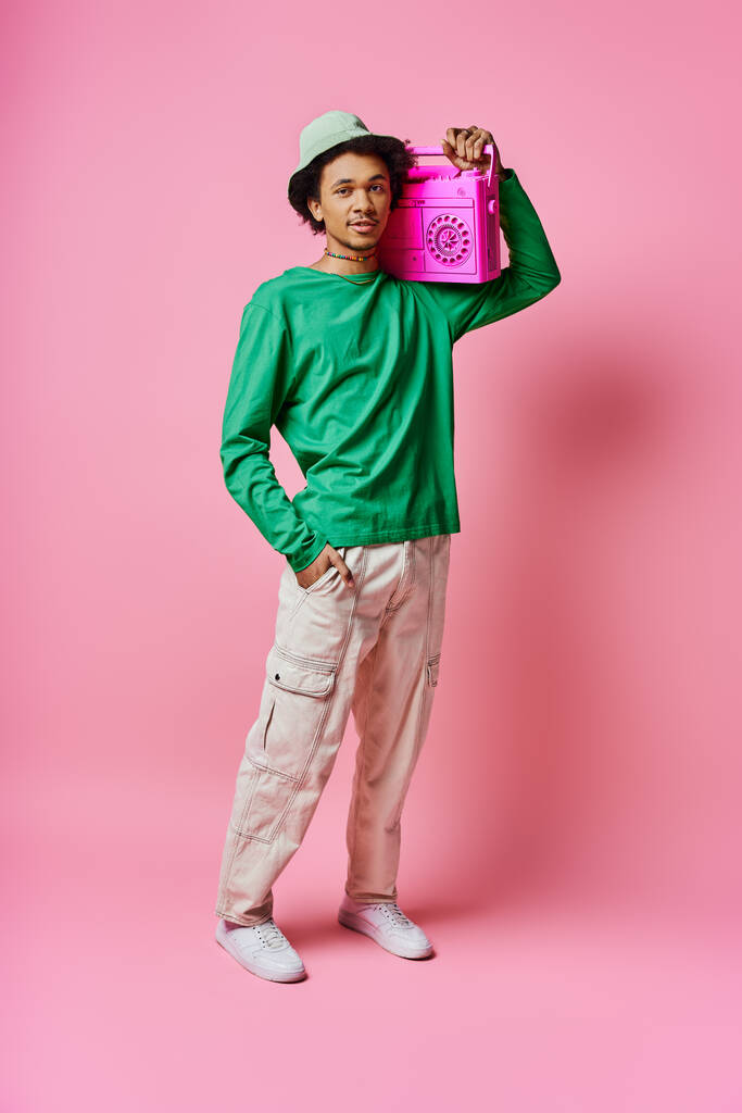 Um jovem afro-americano alegre com uma camisa verde segura uma boombox rosa, mostrando emoção em um fundo rosa. - Foto, Imagem