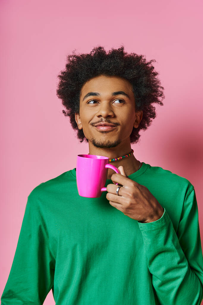 Ένας χαρούμενος, νεαρός Αφρο-Αμερικάνος με περιστασιακή ενδυμασία κρατάει ένα φλιτζάνι μπροστά στο πρόσωπό του με ροζ φόντο.. - Φωτογραφία, εικόνα