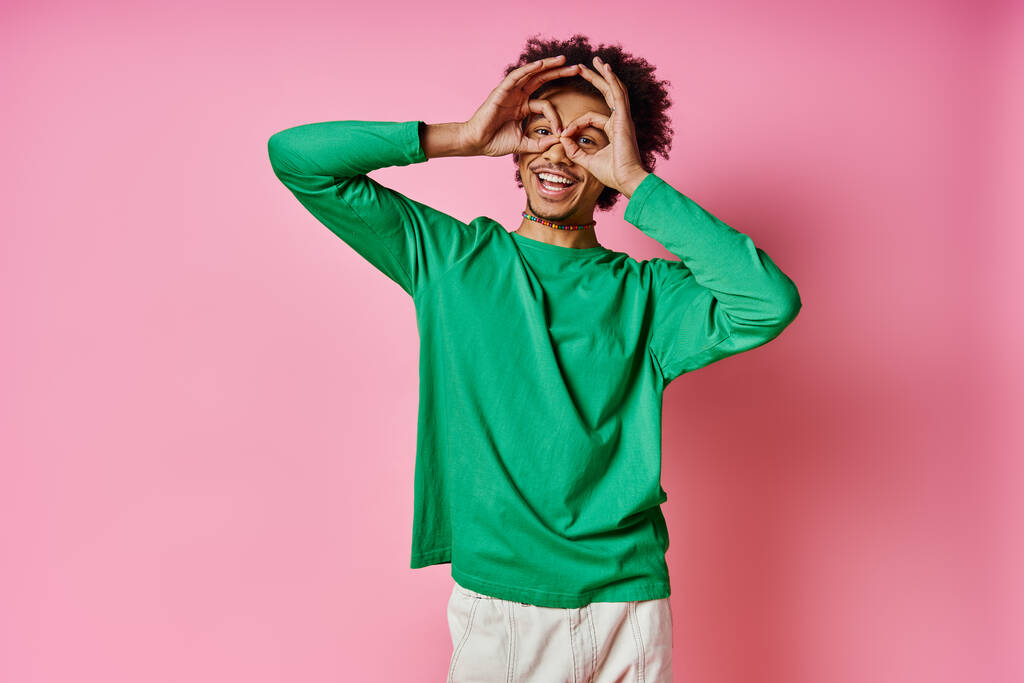 Χαρούμενος νεαρός Αφροαμερικάνος με πράσινο πουκάμισο που καλύπτει τα μάτια του, εκφράζοντας χαρούμενη συγκίνηση σε ροζ φόντο. - Φωτογραφία, εικόνα