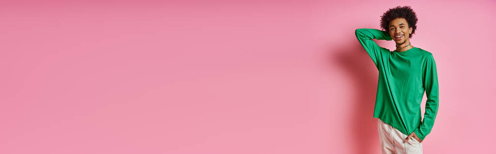 Ο κατσαρομάλλης Αφροαμερικανός με πράσινο πουκάμισο ακουμπά στον λαμπερό ροζ τοίχο, αποπνέοντας συναίσθημα και ηρεμία. - Φωτογραφία, εικόνα