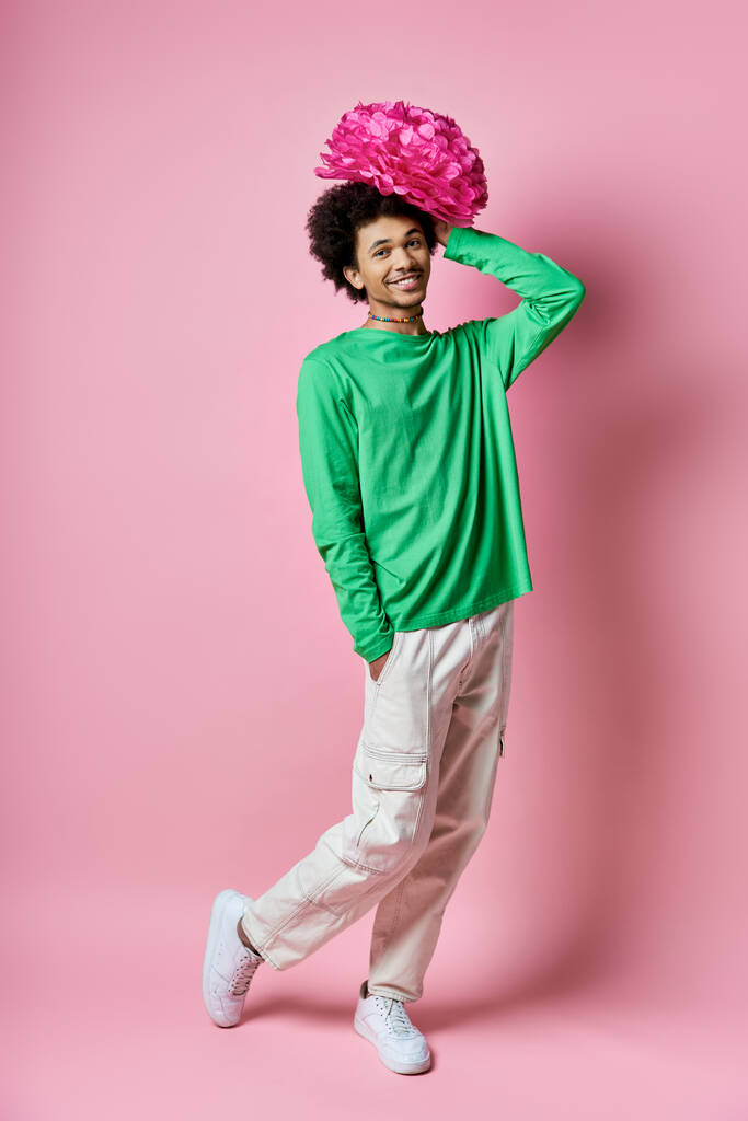 Iloinen nuori afroamerikkalainen mies vihreässä paidassa ja valkoisissa housuissa vaaleanpunainen kukka päässä vaaleanpunaista taustaa vasten. - Valokuva, kuva