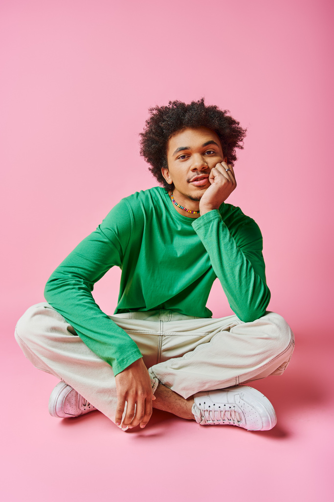mladý afroameričan s kudrnatými vlasy na sobě zelené tričko a bílé kalhoty na růžovém pozadí. - Fotografie, Obrázek