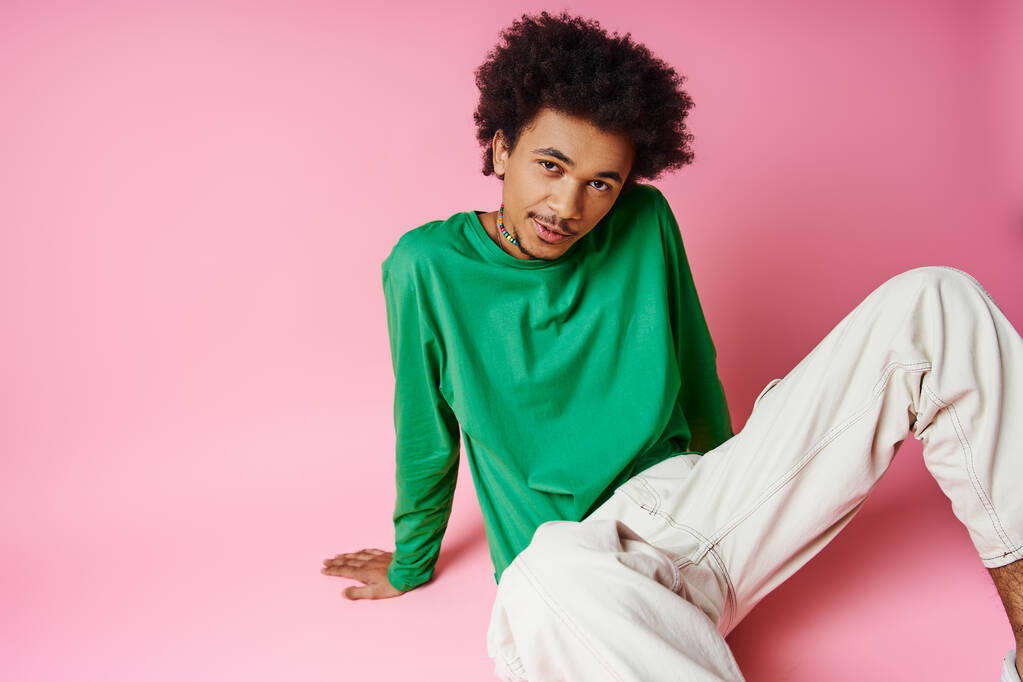 nuori afroamerikkalainen mies, jolla on kiharat hiukset yllään rento vihreä paita ja valkoiset housut vaaleanpunaisella taustalla. - Valokuva, kuva