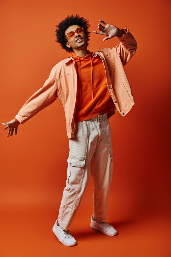 Um jovem afro-americano moderno com cabelo encaracolado posa confiantemente em camisa e calças brancas contra um fundo laranja vibrante. - Foto, Imagem
