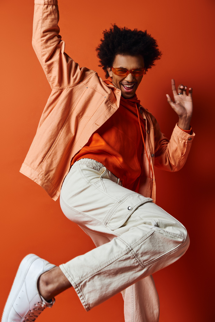 Curly African American man in trendy outfit and sunglasses, jumping energetically in the air against orange background. - Photo, Image