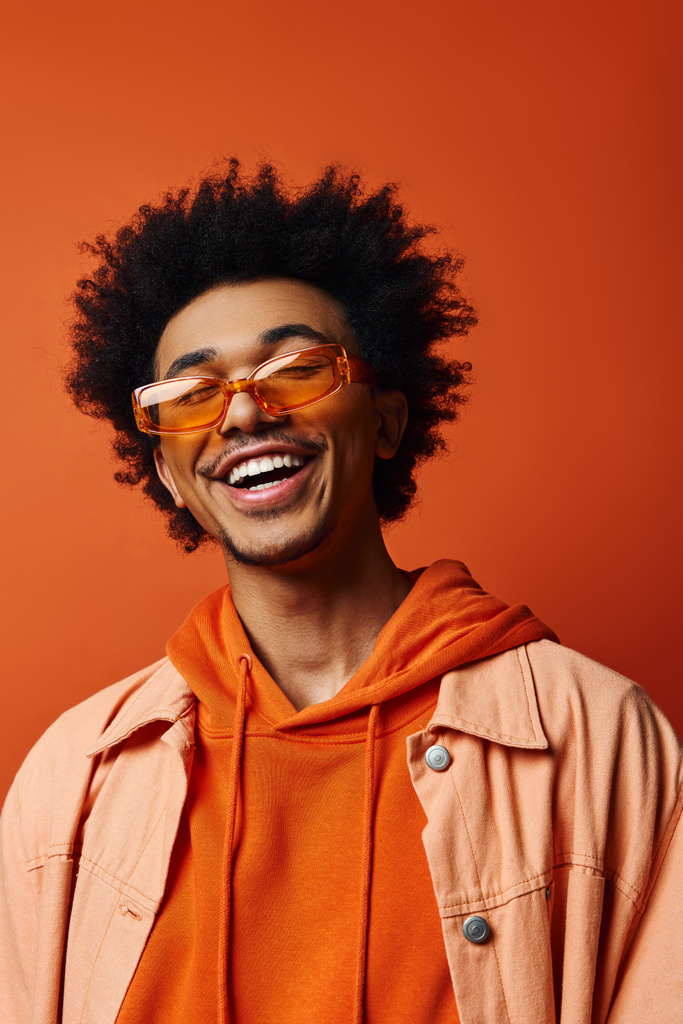 Elegante giovane uomo afro-americano con cappuccio arancione e occhiali da sole con capelli afro ricci su uno sfondo arancione vibrante, esprimendo emozioni. - Foto, immagini