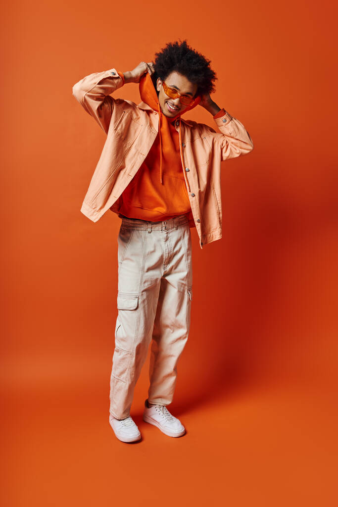 Uomo afroamericano riccio in camicia arancione alla moda e pantaloni kaki, stile trasudante e carisma su uno sfondo arancione audace. - Foto, immagini