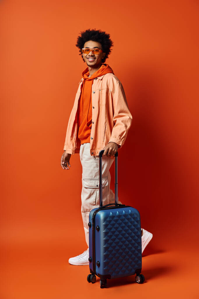 Un jeune homme afro-américain élégant debout avec une valise devant un mur orange, respirant la confiance et le charisme. - Photo, image