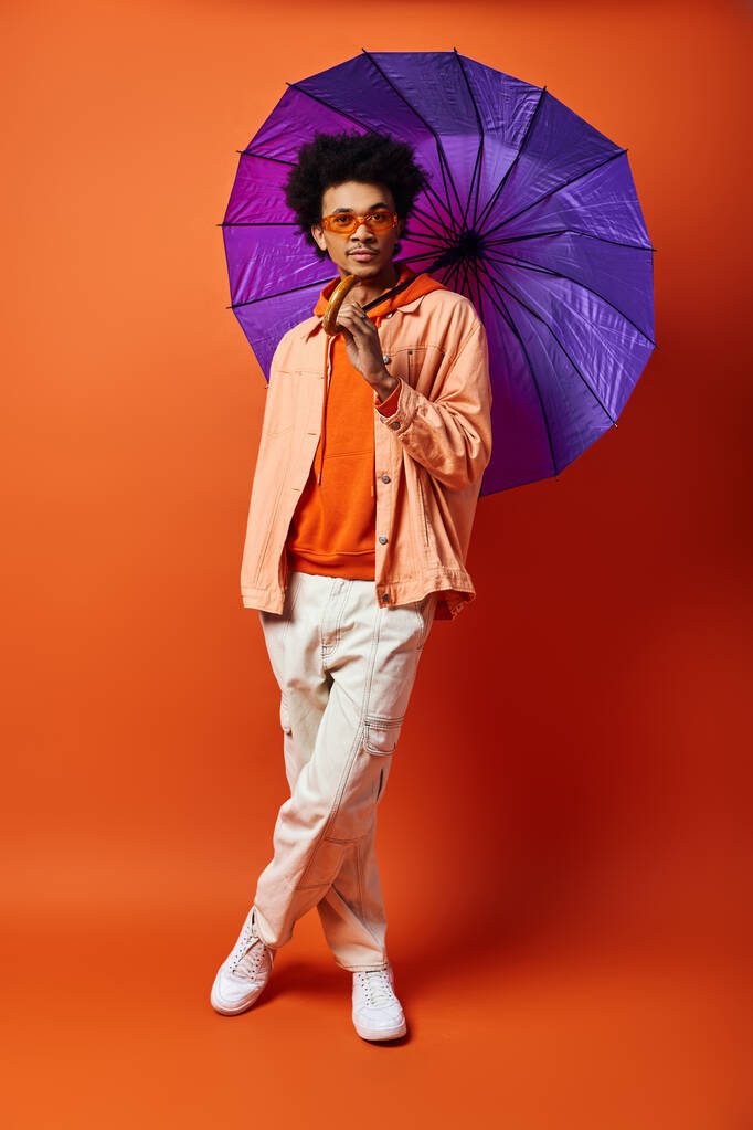 A young African American man with curly hair holds a purple umbrella against a bold orange backdrop, exuding style and personality. - Photo, Image