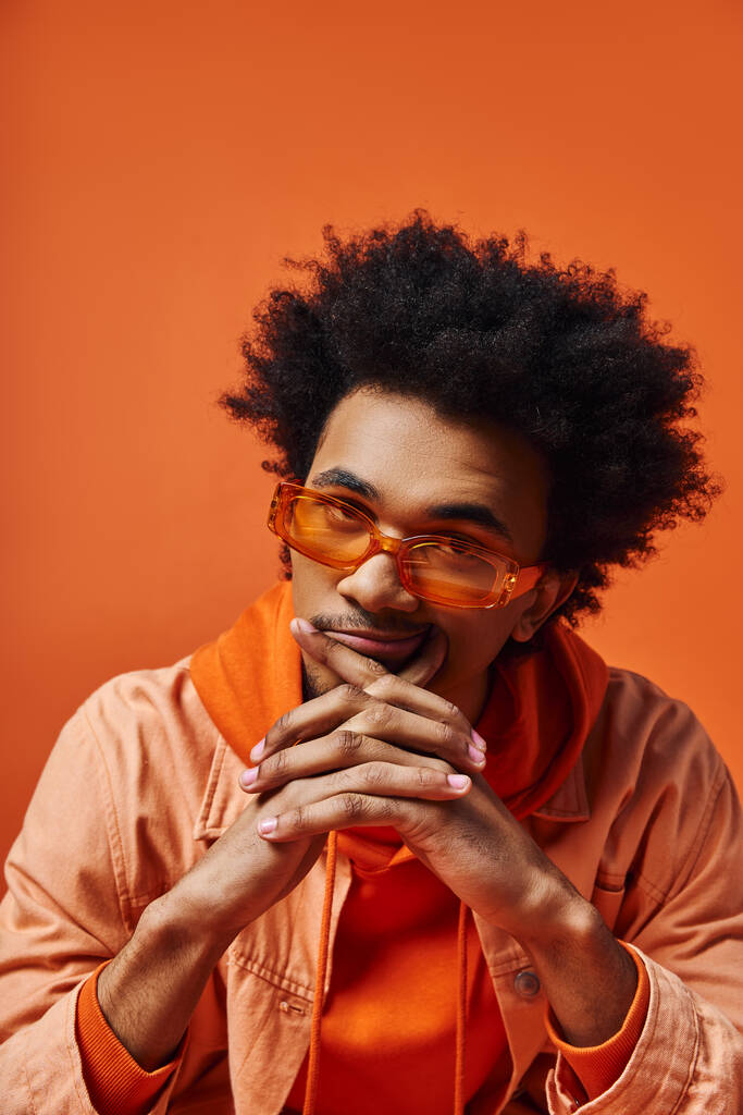 Модний молодий афроамериканський чоловік з кучерявим волоссям в окулярах і апельсиновою курткою на яскравому помаранчевому фоні. - Фото, зображення