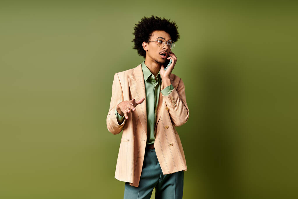 鮮やかな緑の背景に対して携帯電話で話すスーツとサングラスのスタイリッシュな若いアフリカ系アメリカ人男性. - 写真・画像