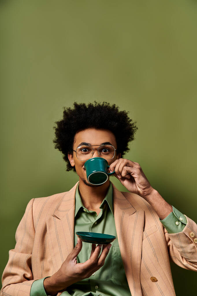 Um jovem afro-americano elegante vestindo trajes da moda e óculos de sol com uma xícara de café em sua boca em um fundo verde. - Foto, Imagem