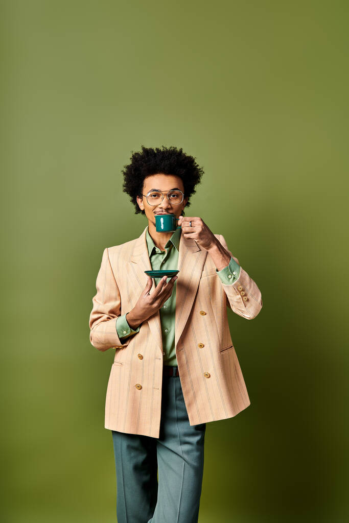 Ein junger afroamerikanischer Mann in modischer Kleidung und Sonnenbrille hält vor grünem Hintergrund eine Tasse Kaffee in der Hand. - Foto, Bild