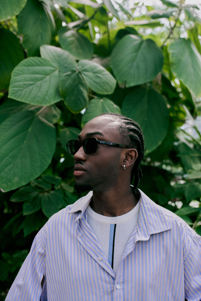 A handsome African American man with sophisticated style stands in front of a lush green tree, wearing sunglasses. - Photo, Image