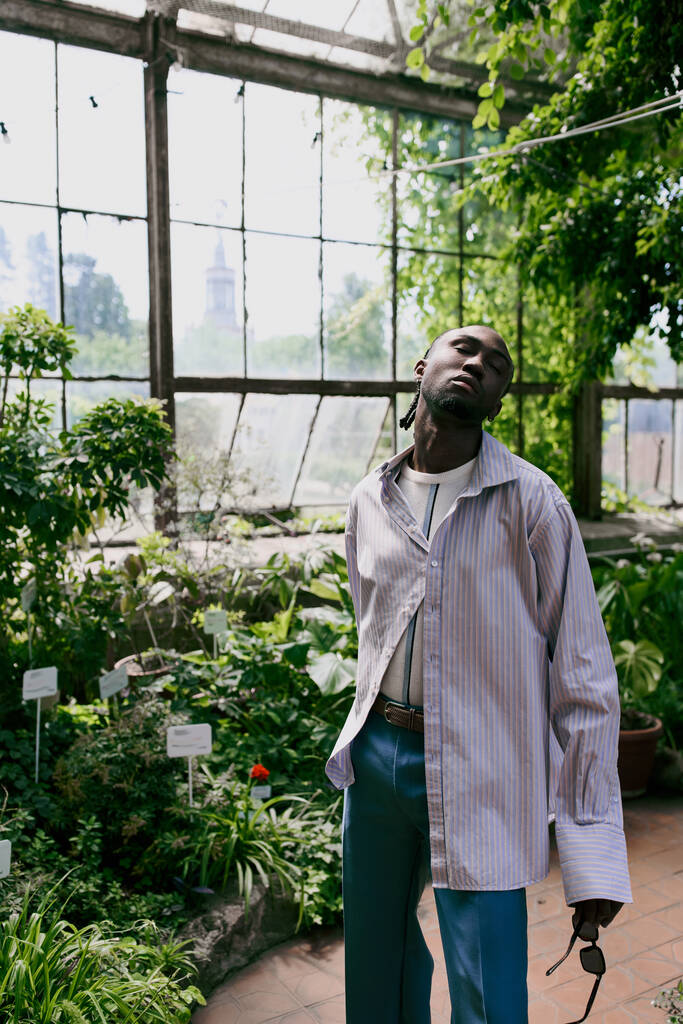 Jóképű afro-amerikai férfi kifinomult öltözékben áll buja zöld üvegházban.. - Fotó, kép