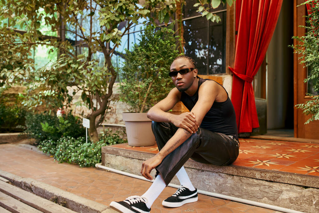 Jóképű afro-amerikai férfi, elegáns stílusban, keresztbe tett lábbal ül egy lépésben egy élénk zöld kertben.. - Fotó, kép