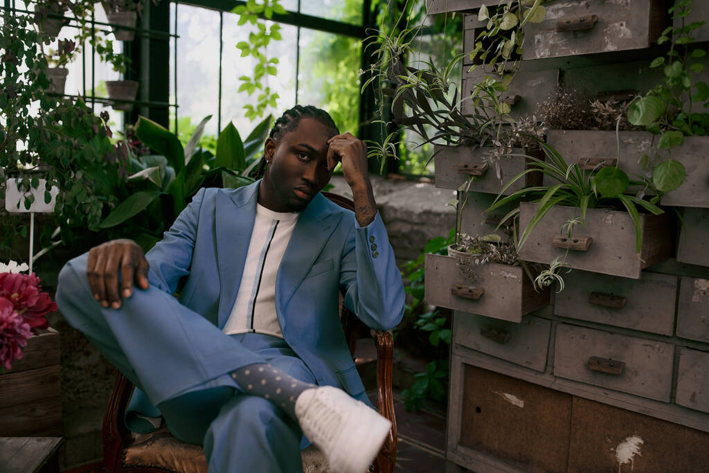 Um homem afro-americano bonito em um terno azul sentado em uma cadeira em um jardim verde vívido. - Foto, Imagem