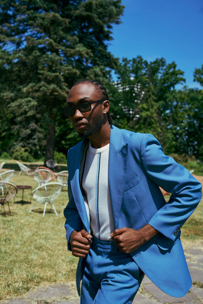 A sophisticated African American man poses in a vivid green garden in a blue suit and sunglasses. - Photo, Image